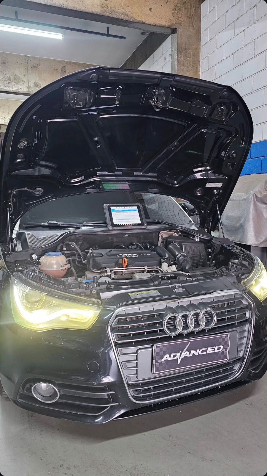 Audi preto com o capô aberto e um tablet apoiado sobre o motor, com a placa decorativa Advanced na grade.