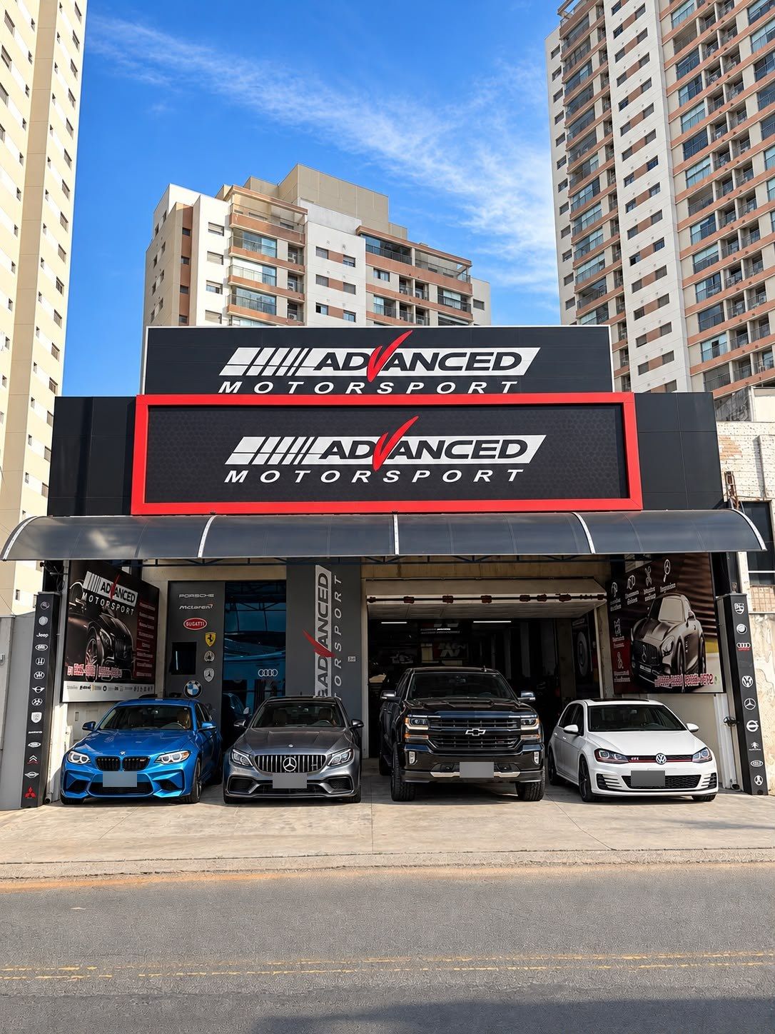 Fachada da Advanced Motorsport, com letreiro preto e vermelho, a entrada da oficina e quatro carros estacionados na frente.