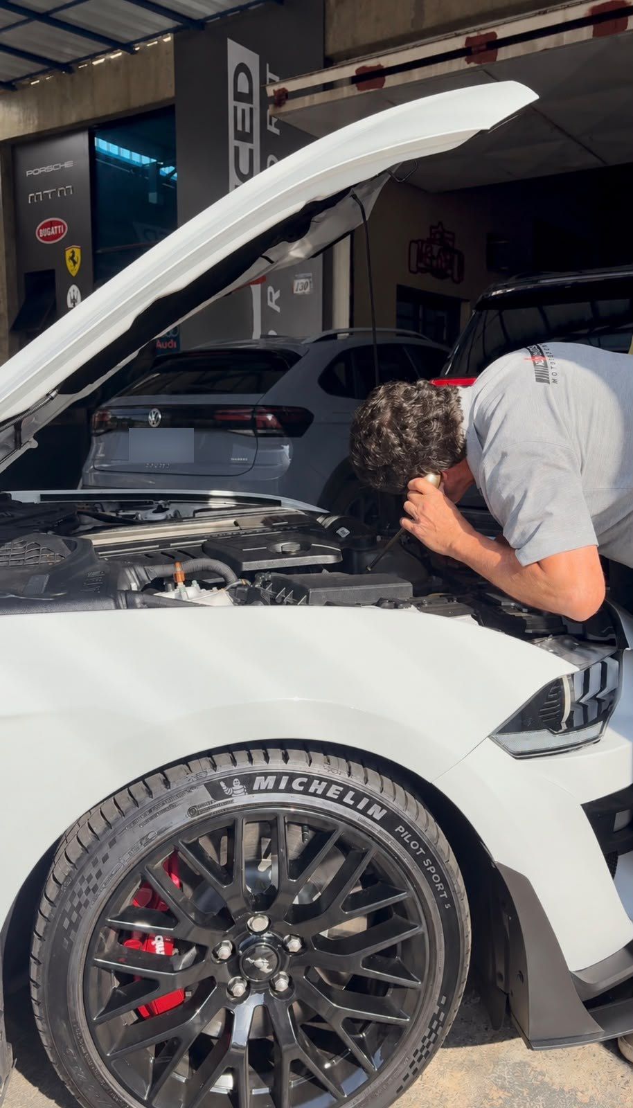 Ricardo Ramos inspecionando o motor de um Mustang branco, com o capô aberto, na entrada da oficina.