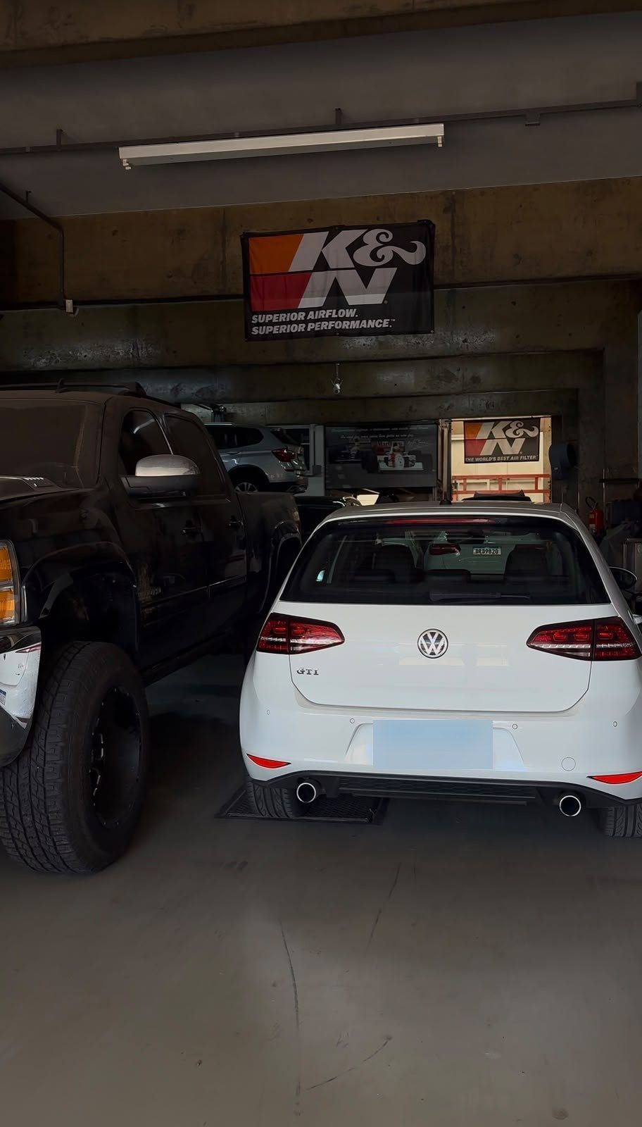 Volkswagen branco visto de trás na oficina, com banner K&N ao fundo.